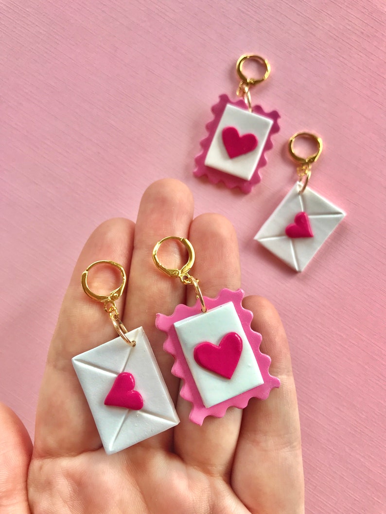 May include: A pair of earrings featuring a pink and white envelope with a pink heart design. The earrings are made of polymer clay and have gold hoops.