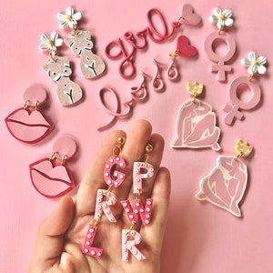 May include: Pink and white polymer clay earrings with various designs, including lips, flowers, letters, and female figures. The earrings are displayed on a pink background.