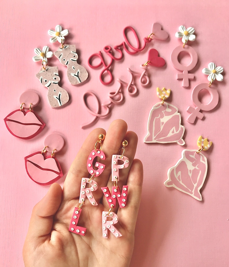 May include: Pink and white polymer clay earrings with various designs, including lips, flowers, letters, and female figures. The earrings are displayed on a pink background.