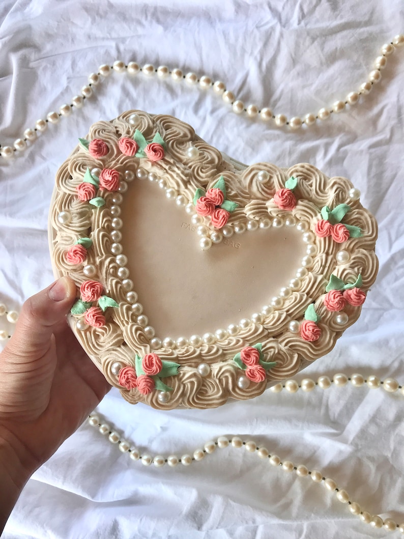 May include: A heart-shaped trinket box with a pearl and floral design. The box is made of white ceramic and features a raised design of pearls, pink roses, and green leaves. The box is decorated with the words "Made in Japan".