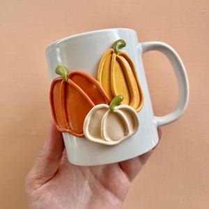 May include: White ceramic mug with three dimensional orange, yellow, and white pumpkins attached to the side.