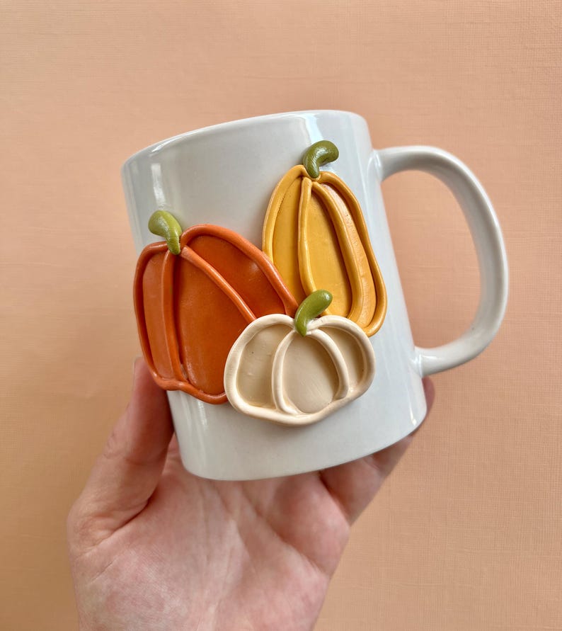 May include: White ceramic mug with three dimensional orange, yellow, and white pumpkins attached to the side.