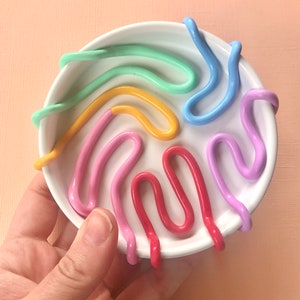 Puede incluir: Un plato de cer&aacute;mica blanca con un dise&ntilde;o colorido. El plato tiene un dise&ntilde;o en relieve de l&iacute;neas onduladas en tonos de rosa, rojo, amarillo, verde y azul.