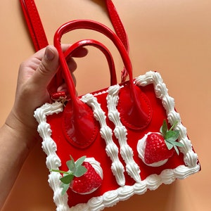 May include: A red faux leather handbag shaped like a strawberry shortcake. The bag has a white cream border and two strawberries on top. The bag has a top handle and a shoulder strap.