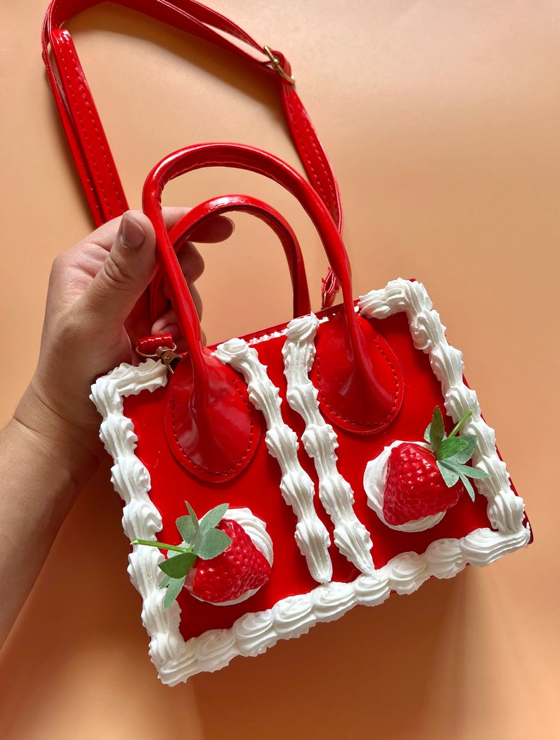 May include: A red faux leather handbag shaped like a strawberry shortcake. The bag has a white cream border and two strawberries on top. The bag has a top handle and a shoulder strap.
