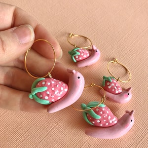 Pu&ograve; includere: Tre paia di orecchini a cerchio dorati con ciondoli a forma di lumaca di fragola rosa e bianca. Le lumache sono rosa con puntini bianchi e foglie verdi.
