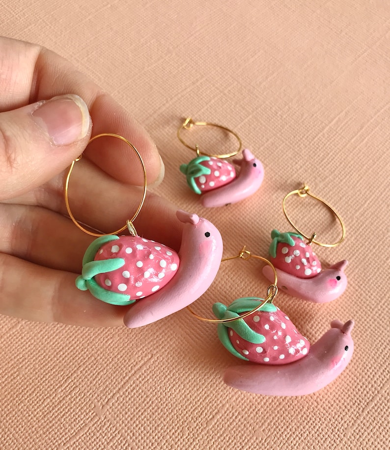 Pu&ograve; includere: Tre paia di orecchini a cerchio dorati con ciondoli a forma di lumaca di fragola rosa e bianca. Le lumache sono rosa con puntini bianchi e foglie verdi.