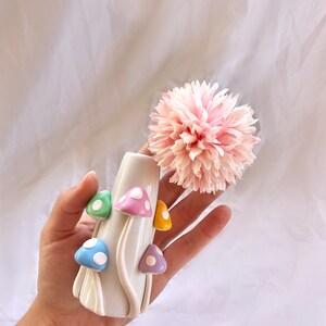 May include: A white ceramic vase shaped like a mushroom with colorful mushroom accents. The vase is holding a pink flower.