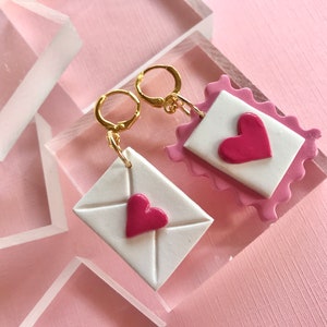 May include: A pair of gold earrings with pink and white clay charms shaped like envelopes with pink hearts. The charms are attached to gold hoops.