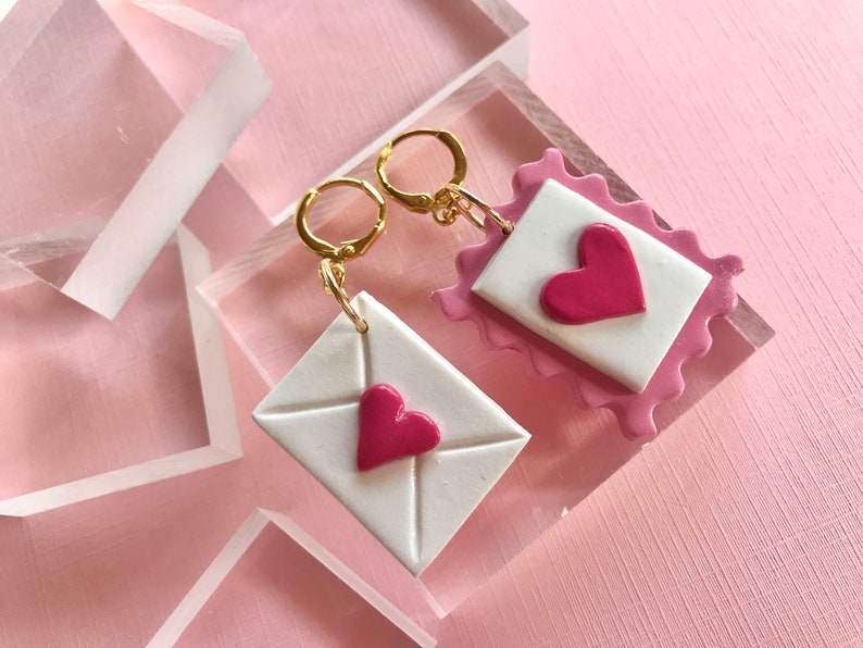 May include: A pair of gold earrings with pink and white clay charms shaped like envelopes with pink hearts. The charms are attached to gold hoops.