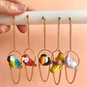 May include: Five pairs of dangle earrings featuring colorful clay bird charms hanging from gold chains. The birds are a blue and yellow parrot, a red parrot, a black and white toucan, a yellow bird with pink accents, and a white cockatoo.