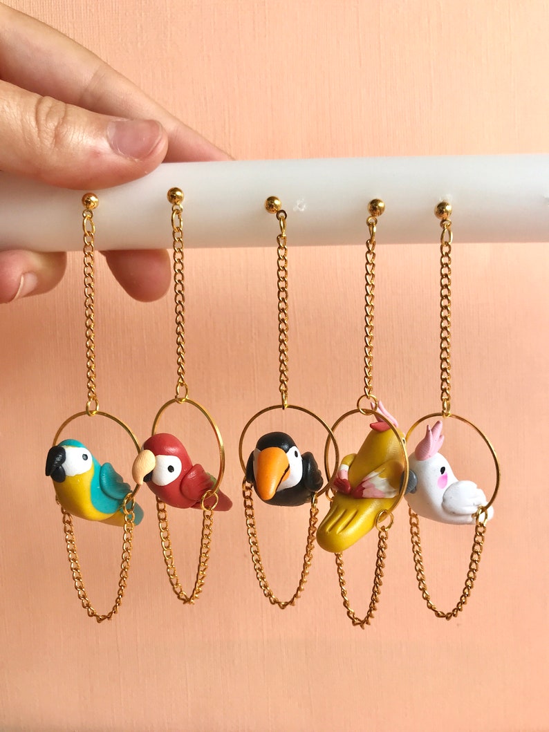 May include: Five pairs of dangle earrings featuring colorful clay bird charms hanging from gold chains. The birds are a blue and yellow parrot, a red parrot, a black and white toucan, a yellow bird with pink accents, and a white cockatoo.