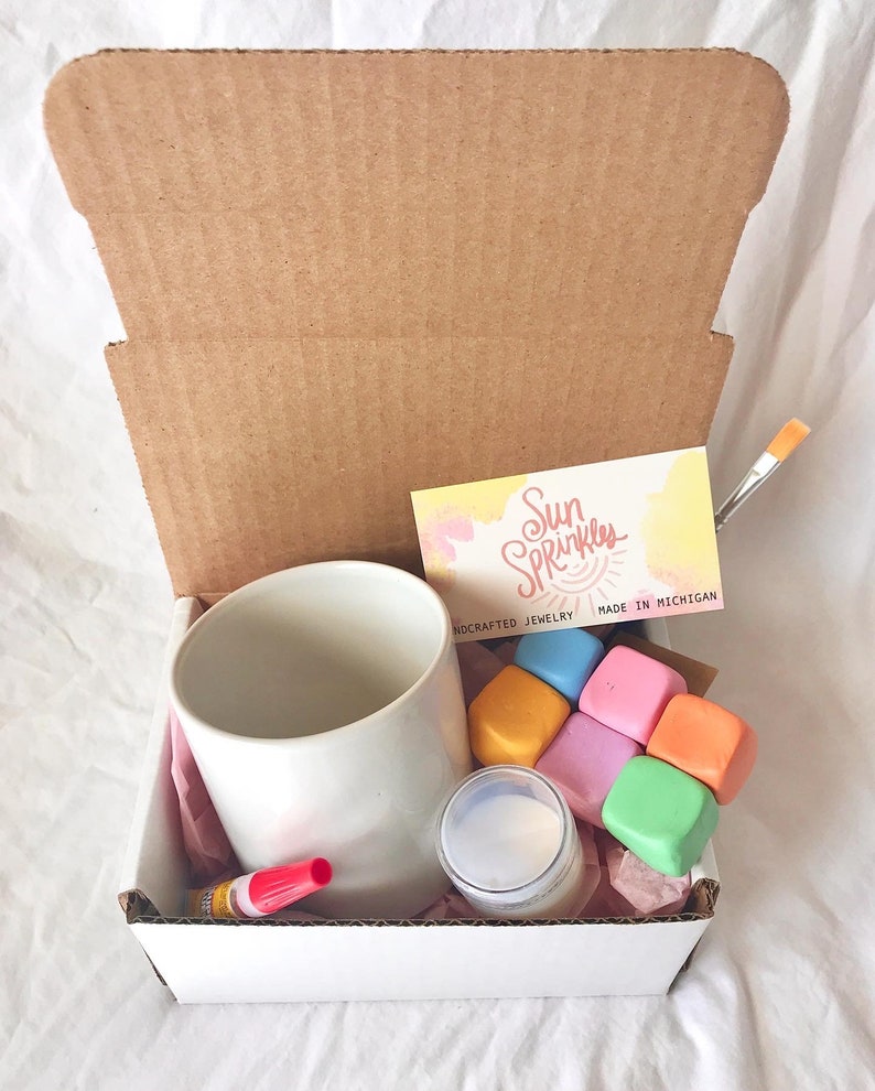 May include: A white ceramic mug, a small jar of white paint, six colorful paint cubes, a red glue stick, a paintbrush, and a business card with the text "Sun Sprinkles Handcrafted Jewelry Made in Michigan".