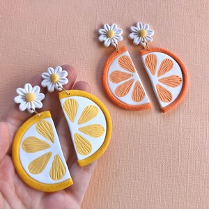 Puede incluir: Pendientes hechos a mano de arcilla polim&eacute;rica con dise&ntilde;o de margarita y rodaja de c&iacute;tricos. Un par es una rodaja de lim&oacute;n con detalles amarillos y blancos, y el otro es una rodaja de naranja con detalles naranjas y blancos. Los pendientes tienen ganchos dorados.