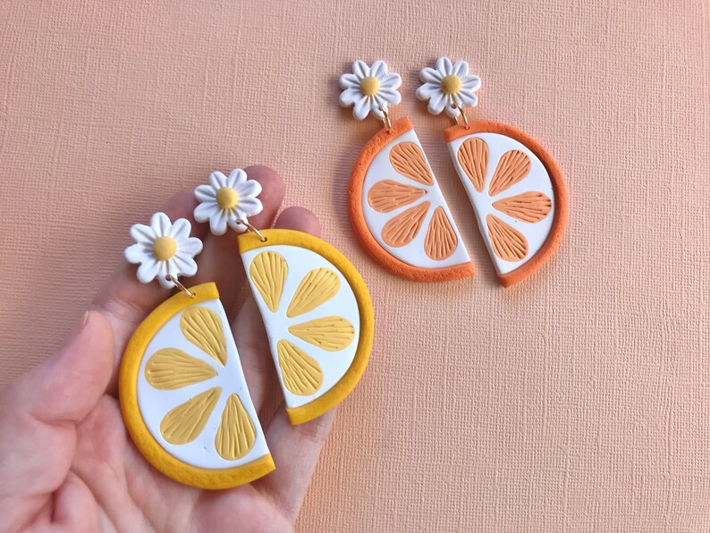 Puede incluir: Pendientes hechos a mano de arcilla polim&eacute;rica con dise&ntilde;o de margarita y rodaja de c&iacute;tricos. Un par es una rodaja de lim&oacute;n con detalles amarillos y blancos, y el otro es una rodaja de naranja con detalles naranjas y blancos. Los pendientes tienen ganchos dorados.
