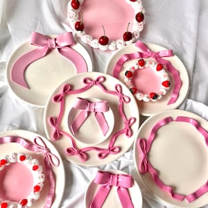 May include: Set of six white ceramic plates with pink ribbon accents. The plates are decorated with pink bows and a pink and white cake design.