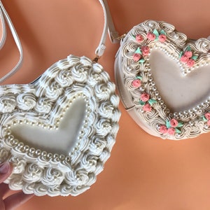 May include: Two white heart-shaped purses with a pearl and floral design. One purse has a white and pink floral design, and the other has a white pearl design.