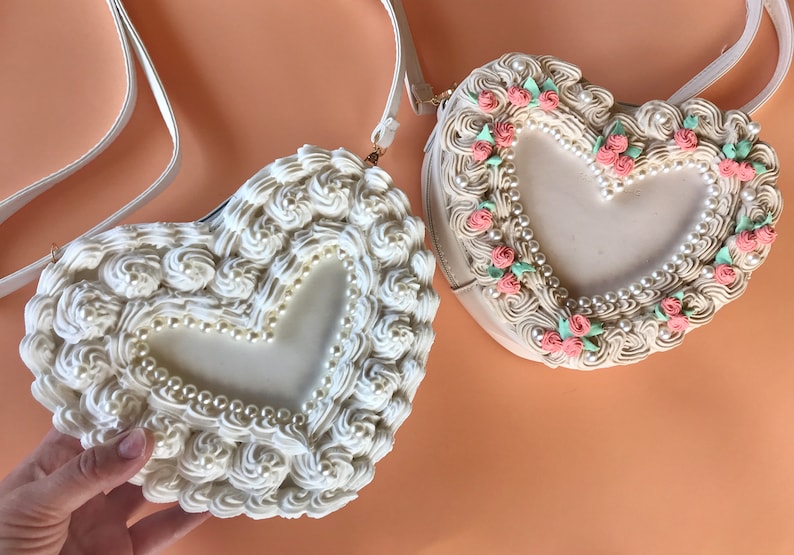 May include: Two white heart-shaped purses with a pearl and floral design. One purse has a white and pink floral design, and the other has a white pearl design.