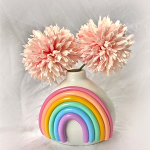 May include: A white ceramic vase with a rainbow design. The vase is filled with two pink flowers.