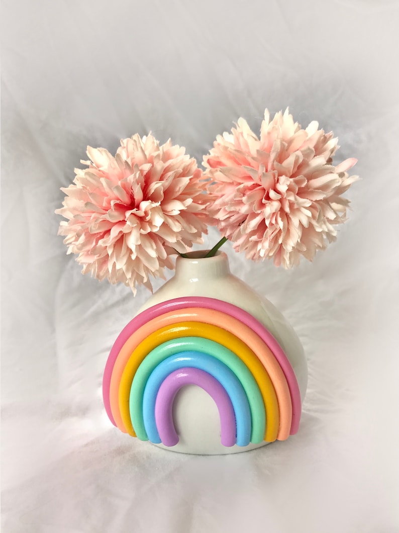 May include: A white ceramic vase with a rainbow design. The vase is filled with two pink flowers.