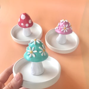 Puede incluir: Tres platos de anillo de cer&aacute;mica con forma de hongo. Los hongos est&aacute;n decorados con puntos blancos, margaritas blancas y margaritas blancas con centros amarillos. Los platos son blancos.