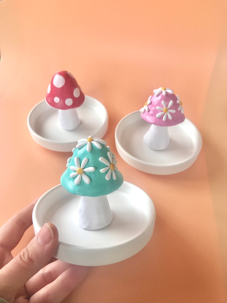 Puede incluir: Tres platos de anillo de cer&aacute;mica con forma de hongo. Los hongos est&aacute;n decorados con puntos blancos, margaritas blancas y margaritas blancas con centros amarillos. Los platos son blancos.