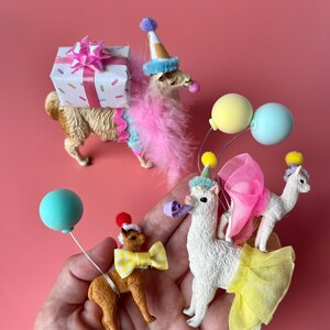K&ouml;nnte beinhalten: Eine Sammlung von Miniatur-Lama-Figuren, die mit festlicher Partykleidung geschm&uuml;ckt sind. Eine tr&auml;gt ein Geschenk, andere tragen Partyh&uuml;te, Schleifen und Luftballons. Die Figuren sind in Braun- und Wei&szlig;t&ouml;nen gehalten, mit bunten Akzenten vor rosa Hintergrund.