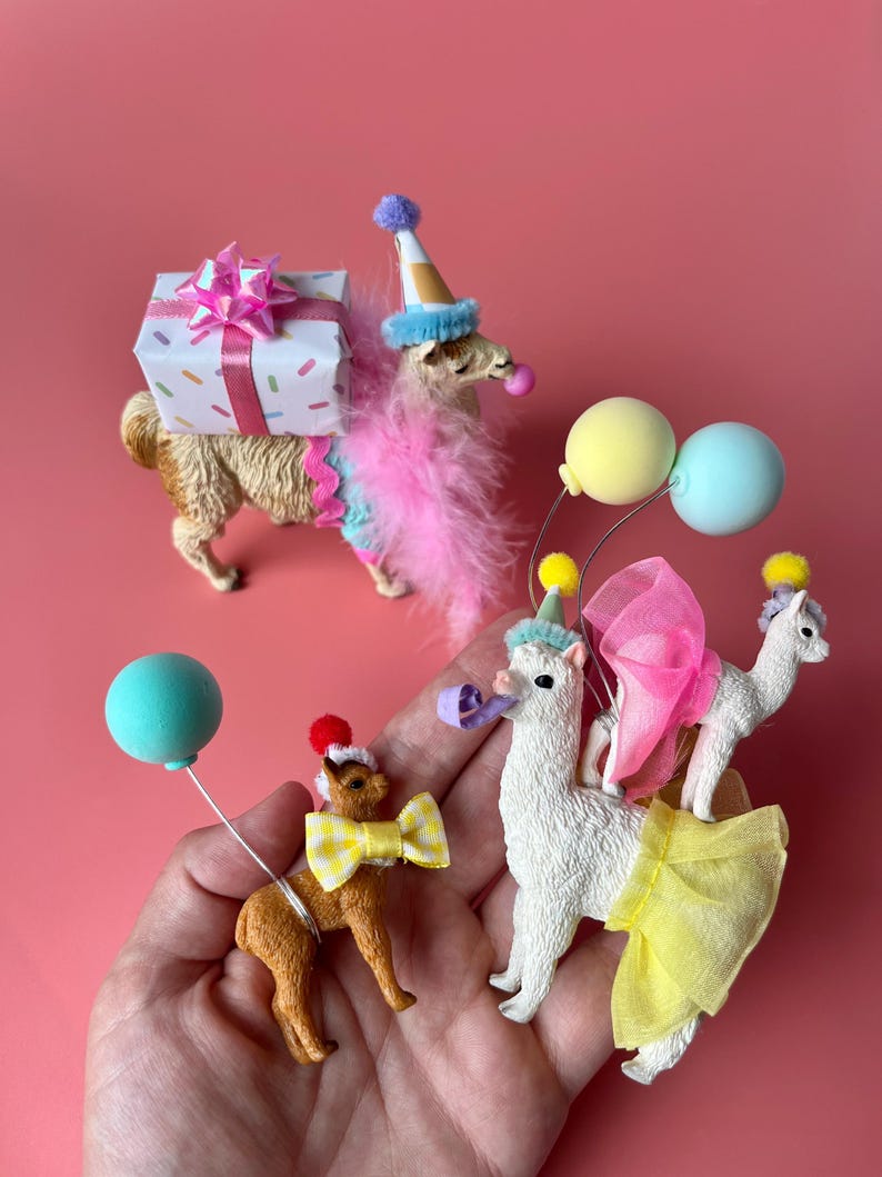 K&ouml;nnte beinhalten: Eine Sammlung von Miniatur-Lama-Figuren, die mit festlicher Partykleidung geschm&uuml;ckt sind. Eine tr&auml;gt ein Geschenk, andere tragen Partyh&uuml;te, Schleifen und Luftballons. Die Figuren sind in Braun- und Wei&szlig;t&ouml;nen gehalten, mit bunten Akzenten vor rosa Hintergrund.