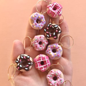 May include: Eight colorful donut-shaped wine glass charms with gold rings. The donuts are decorated with pink, purple, white, and brown frosting and sprinkles.
