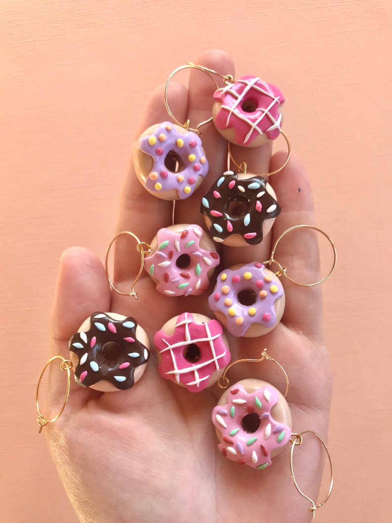 May include: Eight colorful donut-shaped wine glass charms with gold rings. The donuts are decorated with pink, purple, white, and brown frosting and sprinkles.