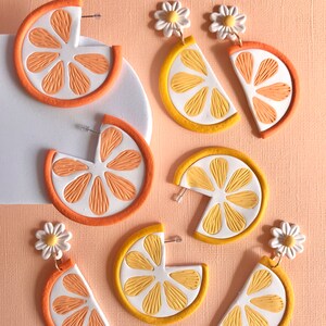 May include: A set of six handmade polymer clay earrings featuring orange and yellow citrus slices with white daisy accents. The earrings are designed to look like half a citrus fruit with a textured surface.