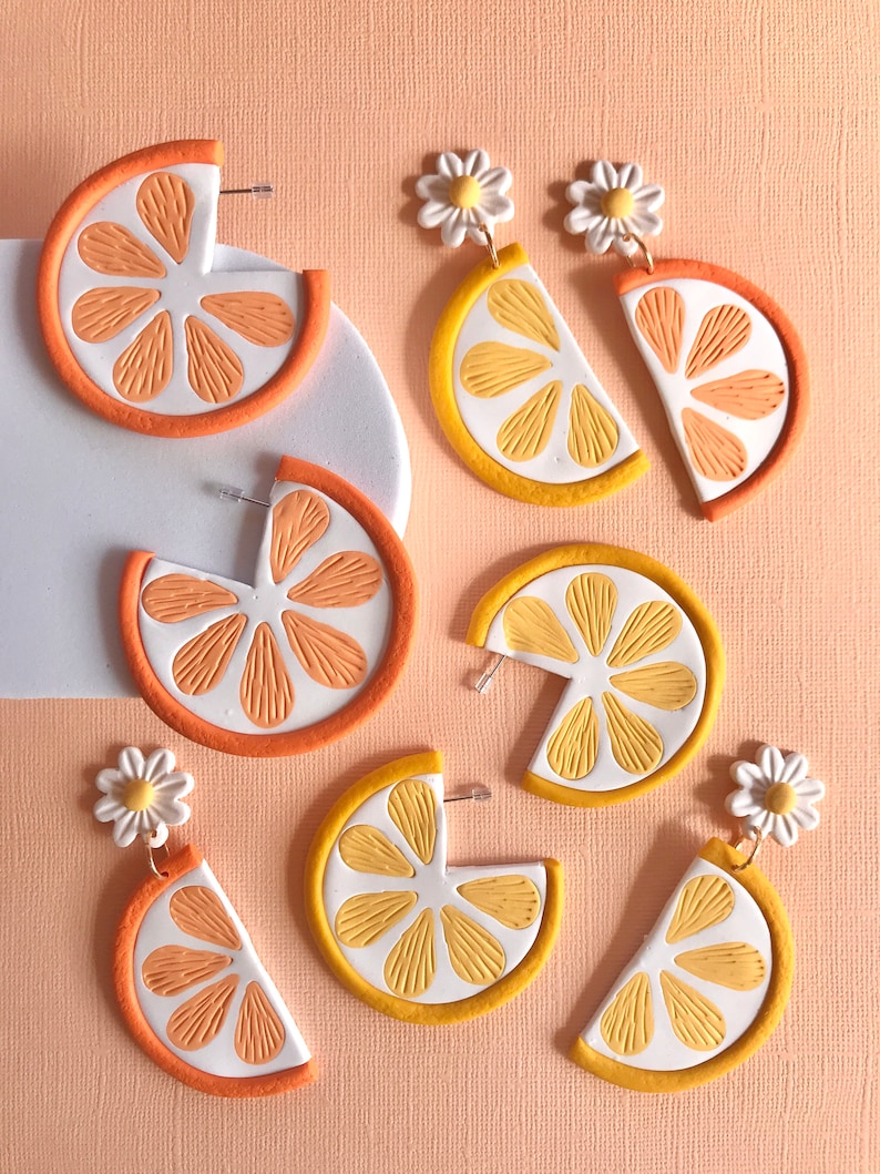 May include: A set of six handmade polymer clay earrings featuring orange and yellow citrus slices with white daisy accents. The earrings are designed to look like half a citrus fruit with a textured surface.