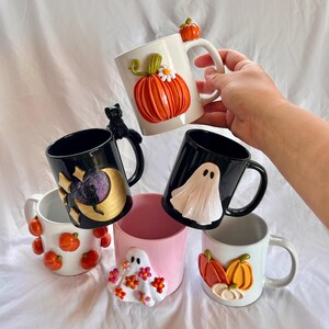 May include: Five Halloween-themed ceramic mugs with different designs. One mug features a pumpkin with a flower, another has a black cat, a third has a ghost, a fourth has a moon and stars, and the last one has a ghost with flowers.