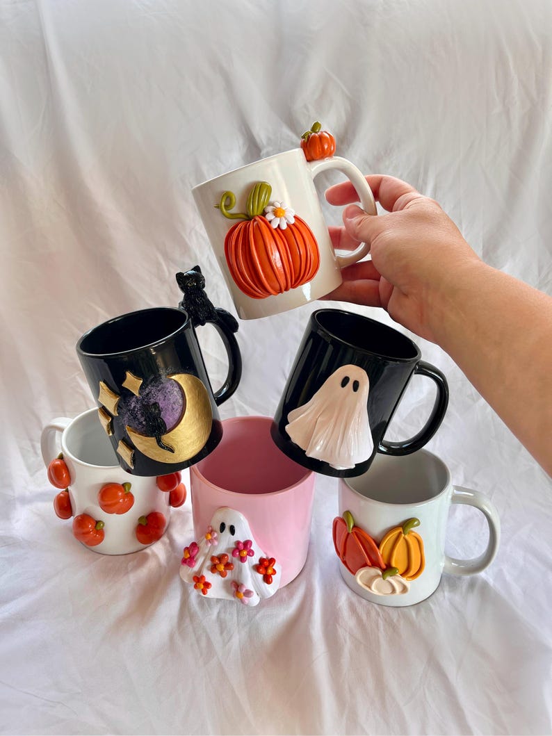 May include: Five Halloween-themed ceramic mugs with different designs. One mug features a pumpkin with a flower, another has a black cat, a third has a ghost, a fourth has a moon and stars, and the last one has a ghost with flowers.