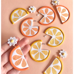 May include: Six pairs of orange slice earrings with a white daisy charm. The earrings are made of polymer clay and have a textured surface.