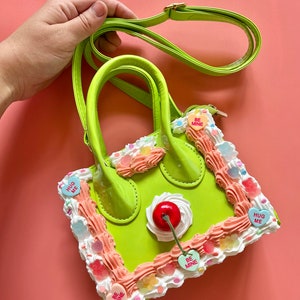 May include: A lime green faux leather handbag shaped like a cake with pink and white frosting, a red cherry on top, and candy hearts that say "Hug Me" and "Be Mine".