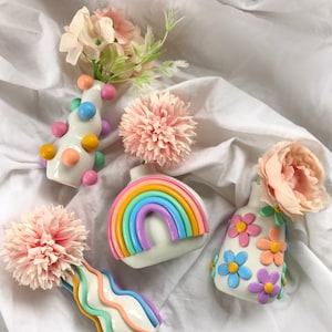May include: Five small ceramic vases with different designs. The vases are white with colorful accents. One vase has a rainbow design, another has a polka dot design, another has a flower design, and two have wavy lines. The vases are on a white fabric background.