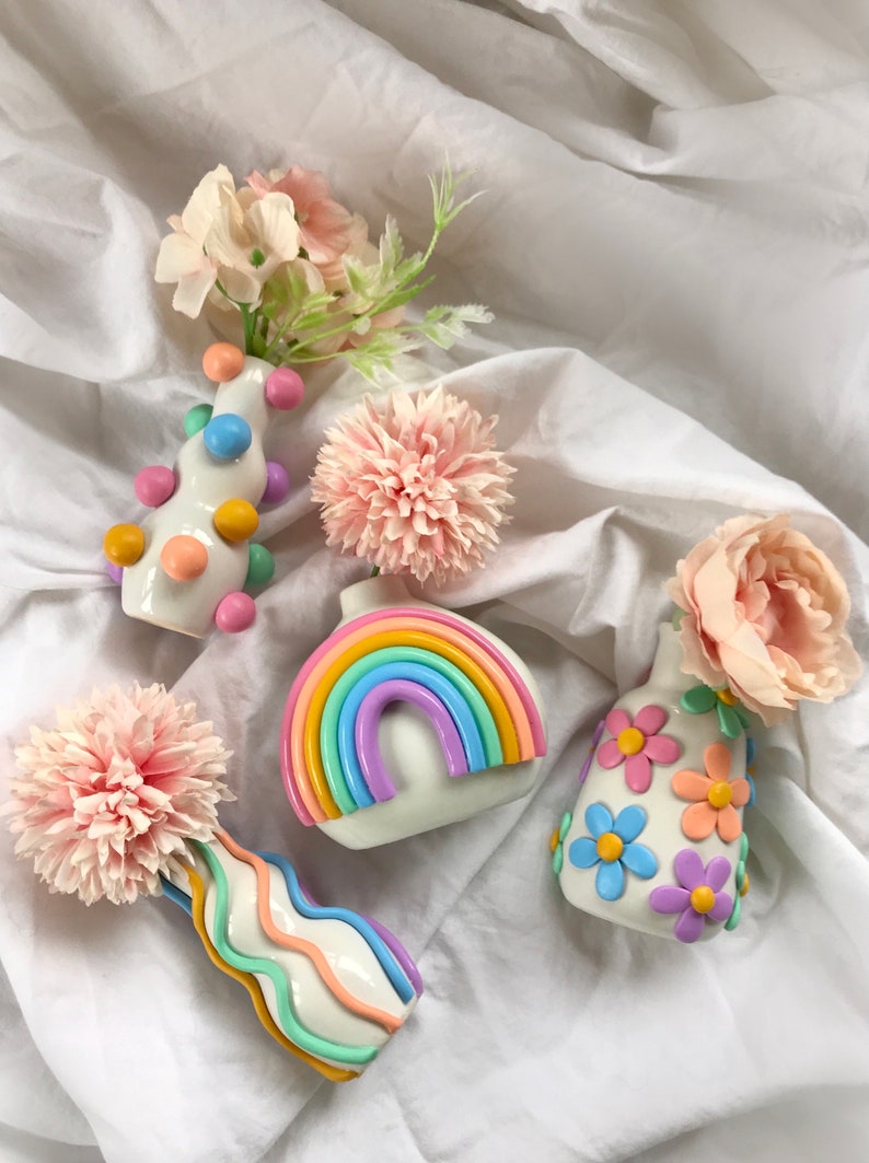 May include: Five small ceramic vases with different designs. The vases are white with colorful accents. One vase has a rainbow design, another has a polka dot design, another has a flower design, and two have wavy lines. The vases are on a white fabric background.