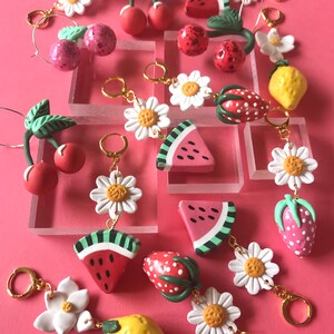 May include: A collection of colorful fruit-themed earrings with gold hoops. The earrings feature cherries, strawberries, lemons, and watermelon slices. Some earrings also have white daisy flowers.