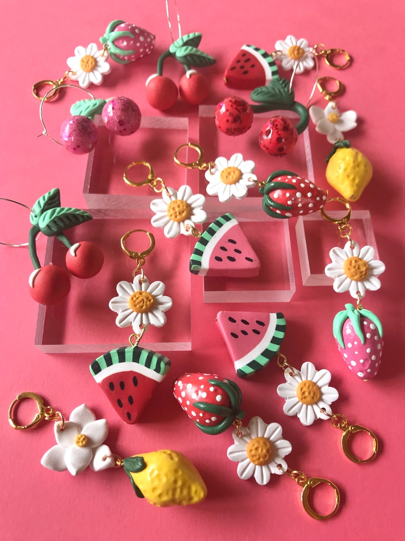 May include: A collection of colorful fruit-themed earrings with gold hoops. The earrings feature cherries, strawberries, lemons, and watermelon slices. Some earrings also have white daisy flowers.