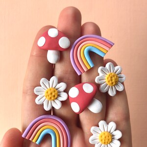 May include: A set of six colorful clay earrings featuring a rainbow, mushroom, and daisy designs. The earrings are handmade and have a whimsical, retro aesthetic.