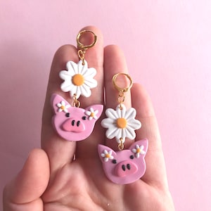 Pu&ograve; includere: Un paio di orecchini pendenti con facce di maiale rosa, fiori di margherita bianchi e cerchietti dorati.