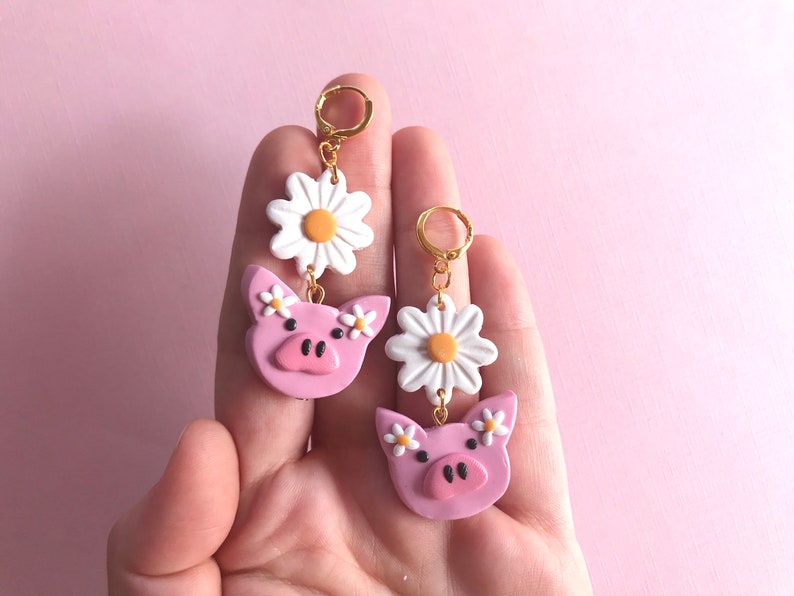 Pu&ograve; includere: Un paio di orecchini pendenti con facce di maiale rosa, fiori di margherita bianchi e cerchietti dorati.