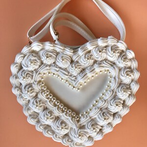 May include: A white heart-shaped handbag with a white pearl trim and a white, ruffled design. The bag has a white strap.