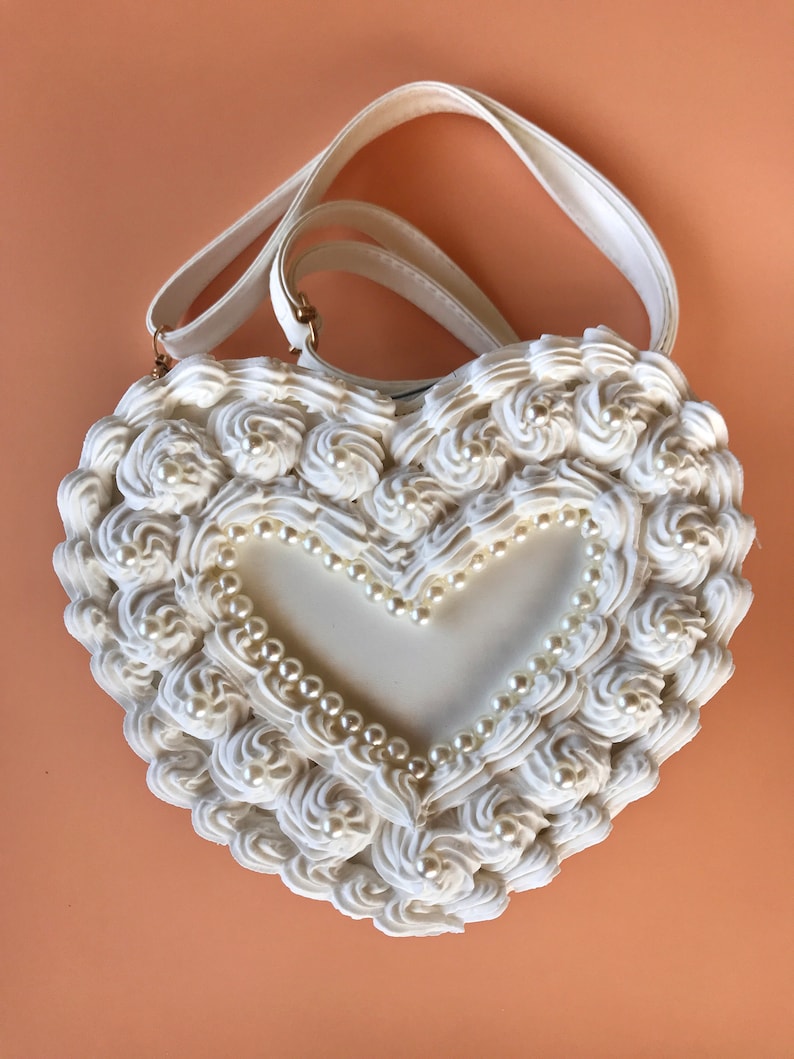 May include: A white heart-shaped handbag with a white pearl trim and a white, ruffled design. The bag has a white strap.