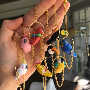 May include: A set of eight colorful bird earrings made of polymer clay. Each bird is attached to a gold chain and hoop. The birds include a pink flamingo, a red cardinal, a black bird, a blue bird, a toucan, a yellow bird, a white cockatoo, and a yellow parakeet.