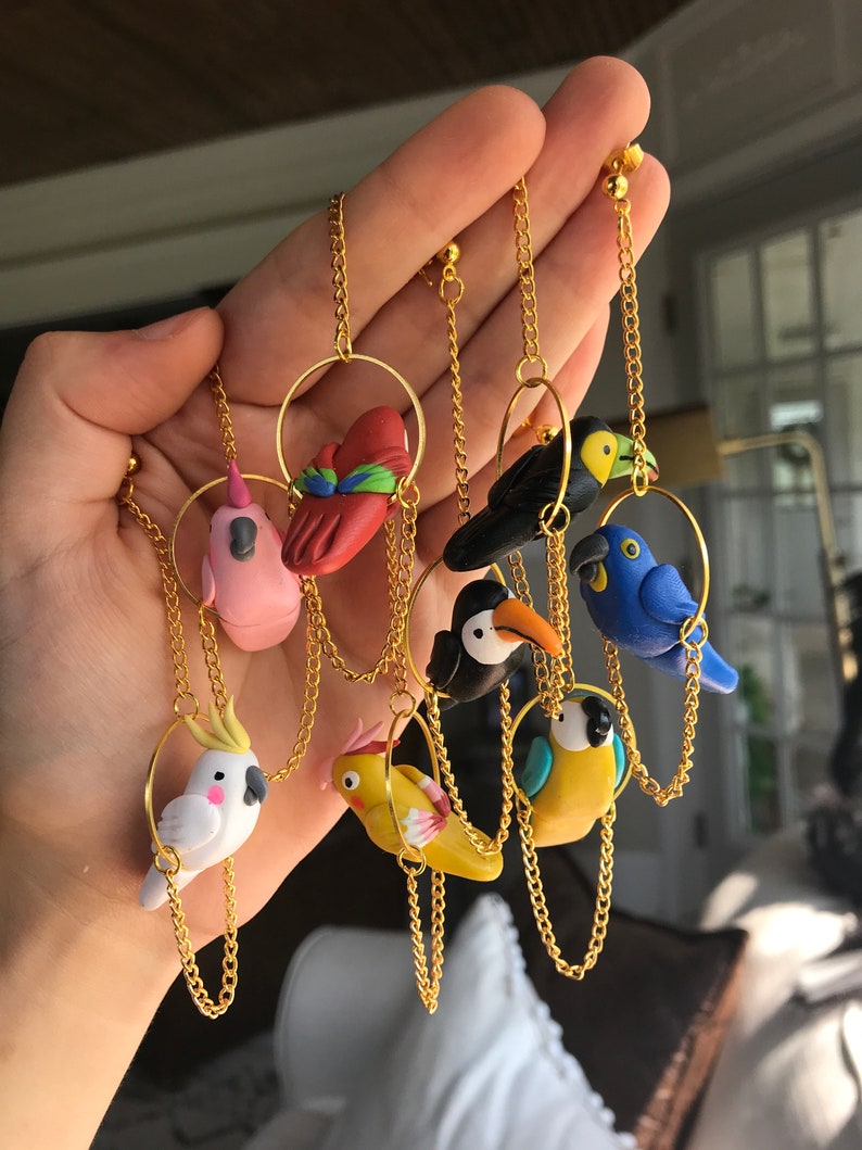 May include: A set of eight colorful bird earrings made of polymer clay. Each bird is attached to a gold chain and hoop. The birds include a pink flamingo, a red cardinal, a black bird, a blue bird, a toucan, a yellow bird, a white cockatoo, and a yellow parakeet.