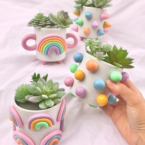 May include: Four small white ceramic planters with different colorful designs. The planters are decorated with rainbows, polka dots, and spikes. Each planter has a small succulent plant inside.