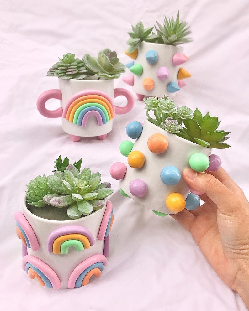 May include: Four small white ceramic planters with different colorful designs. The planters are decorated with rainbows, polka dots, and spikes. Each planter has a small succulent plant inside.