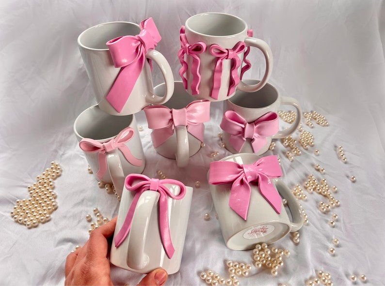 Pu&ograve; includere: Sei tazze in ceramica bianca con fiocchi rosa. Le tazze sono disposte in cerchio su una superficie bianca. I fiocchi sono realizzati in un materiale morbido e malleabile e sono fissati alle tazze con un adesivo resistente.